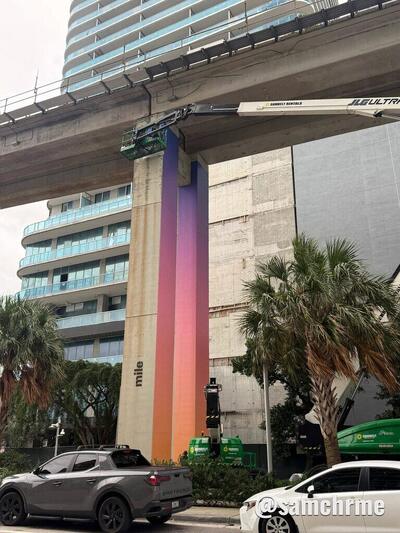 Pilar do metrô de Miami envelopado com as cores de GTA 6 e o cesto de uma plataforma elevatória no topo, com carros estacionados na rua