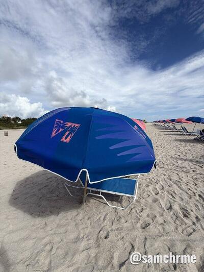 Guarda-sol azul aberto com o logotipo VI rosa, na areia
