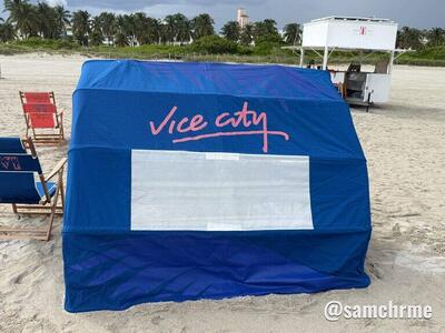 Cabana azul de praia com "Vice City" em rosa e janela branca, com cabine de salva-vidas ao fundo