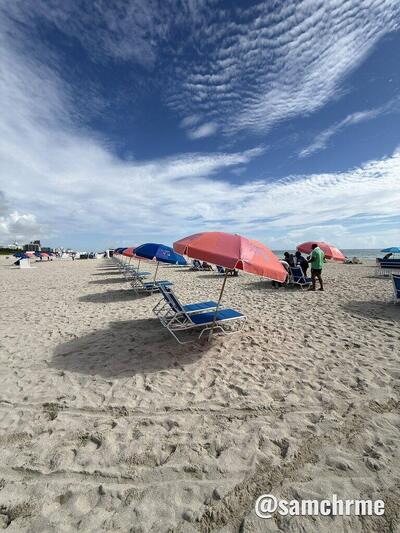 Fileira de guarda-sóis vermelhos e cadeiras azuis na areia de Miami Beach, sob um céu de nuvens