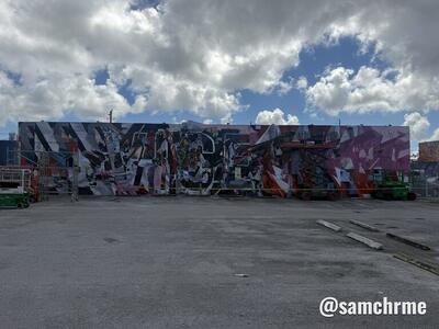 Muro grande com mural abstrato em preto, vermelho e rosa, visto de um estacionamento vazio sob nuvens