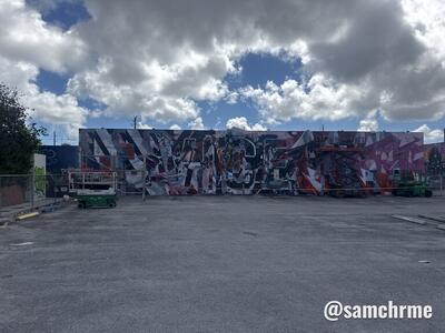 Muro com o mural abstrato em preto, vermelho e rosa, visto de longe, de um estacionamento