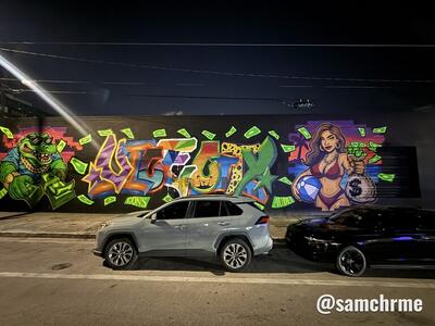 Mural com letras de grafite coloridas ao centro, jacaré à esquerda e mulher de biquíni com saco de dinheiro à direita, à noite, com carros estacionados