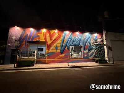 O mural "We ♥ Vice City" à noite, iluminado por refletores, com plataformas elevatórias no passeio